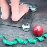 Green measure tape, glass weighing scale, red apple and feet on a scale on a rustic surface. Weight management and healthy wellbeing concept