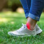 Close-up of a woman's ankle in pain while wearing white sneakers and jeans, standing on grass.