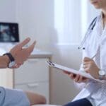 Doctor woman is taking notes on a clipboard while a male patient describes his symptoms in clinic